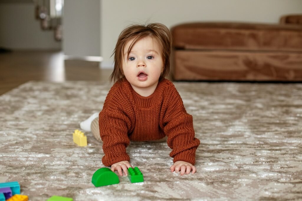 Cute baby girl crawling on floor at home, playing with colored building blocks. Brown sweater.